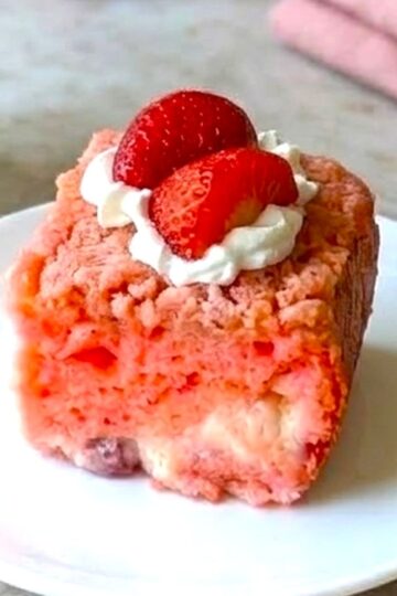 A slice of pink Strawberry Earthquake Cake on a white plate, topped with a dollop of whipped cream and a fresh strawberry half, with a fork nearby.