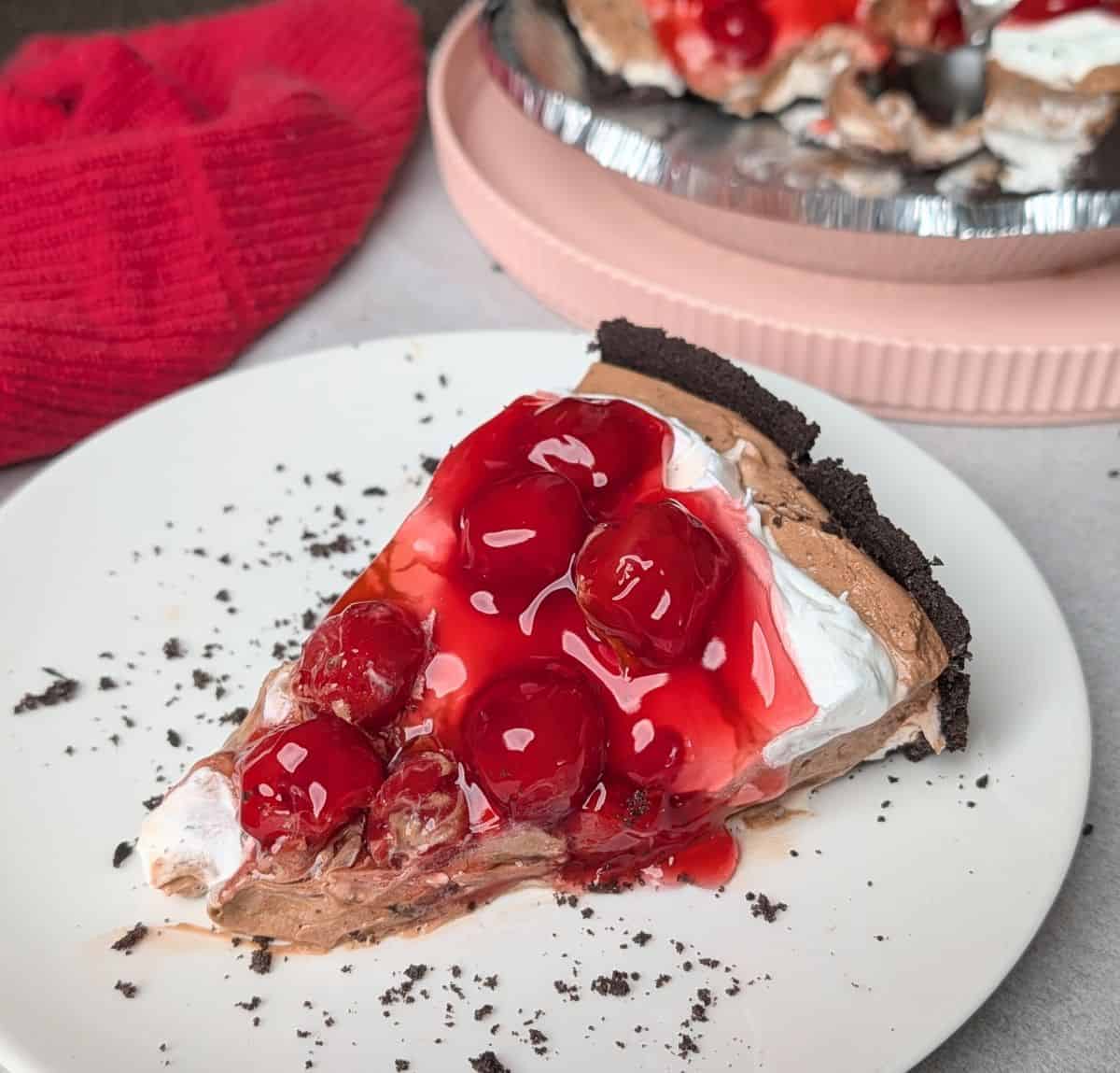 A side view of a sliced Black Forest pie on a white plate, showcasing the chocolate cookie crust, creamy chocolate filling, fluffy whipped cream, and cherry topping.