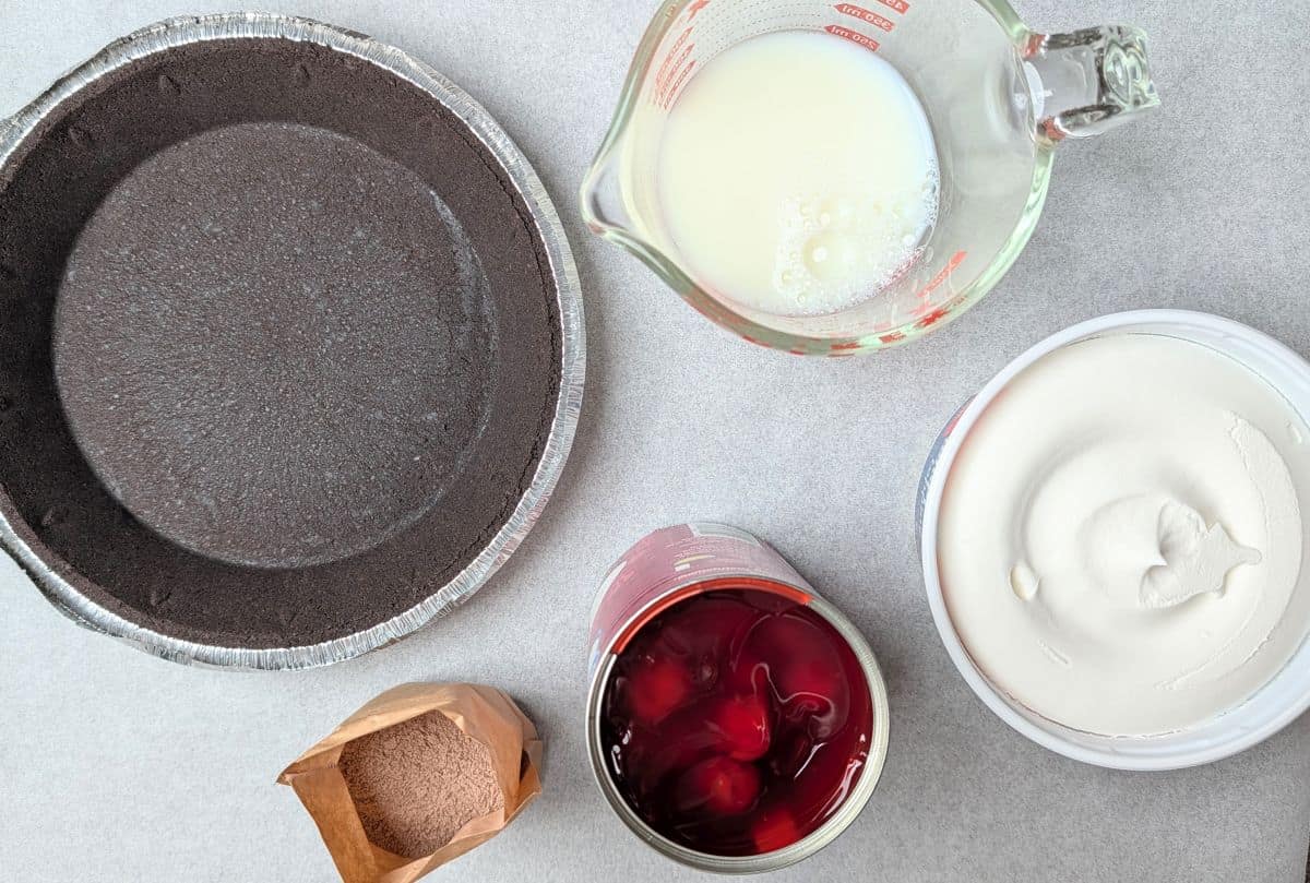 Top-down view of the simple ingredients needed to make an easy Black Forest pie, including a pre-made chocolate cookie crust, milk, and filling components.