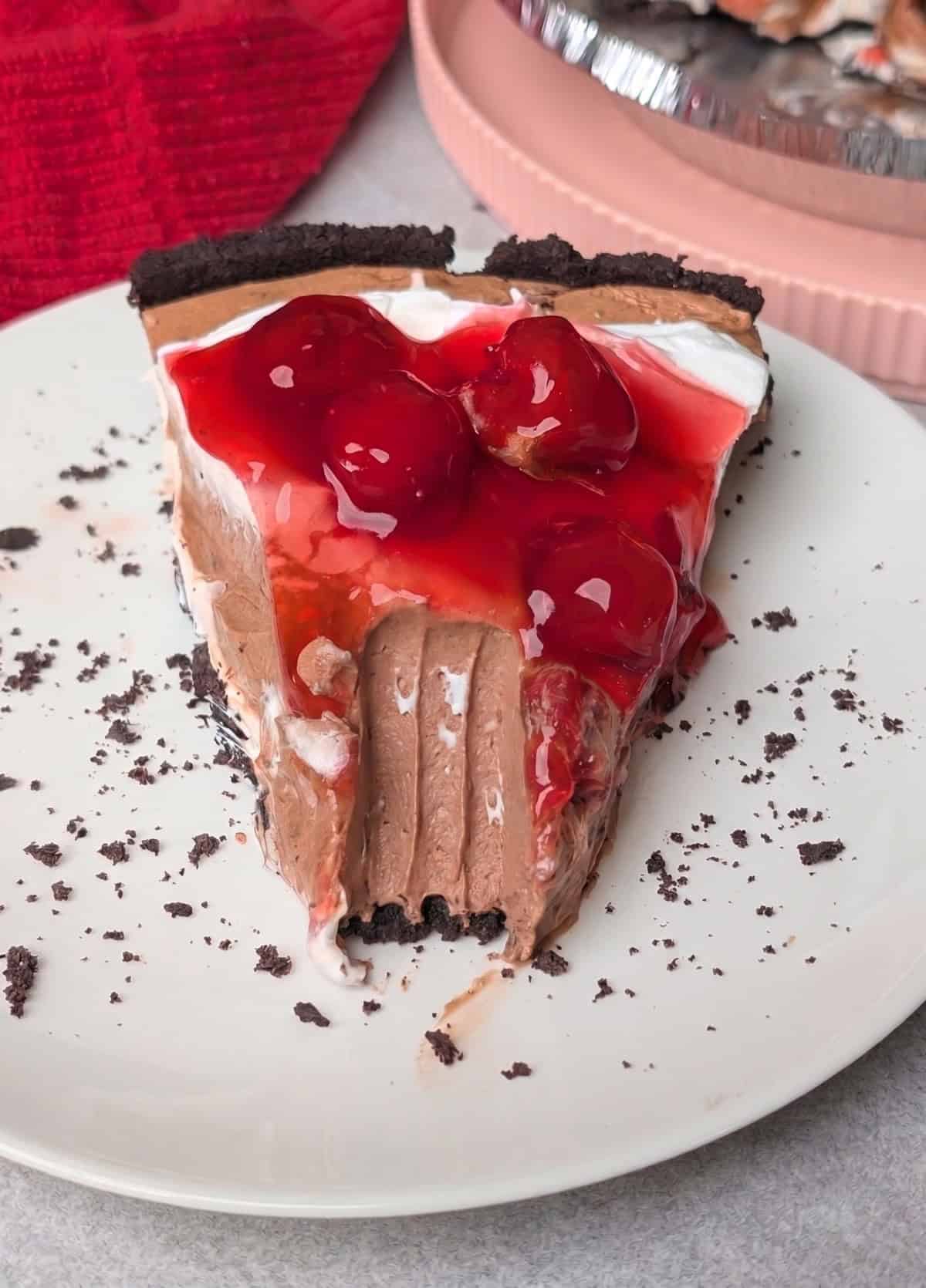 A slice of Black Forest pie with a chocolate crust, creamy chocolate filling, whipped cream, and cherry topping on a white plate with a bite taken out of it.