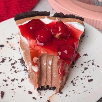A slice of Black Forest pie with a chocolate crust, creamy chocolate filling, whipped cream, and cherry topping on a white plate with a bite taken out of it.