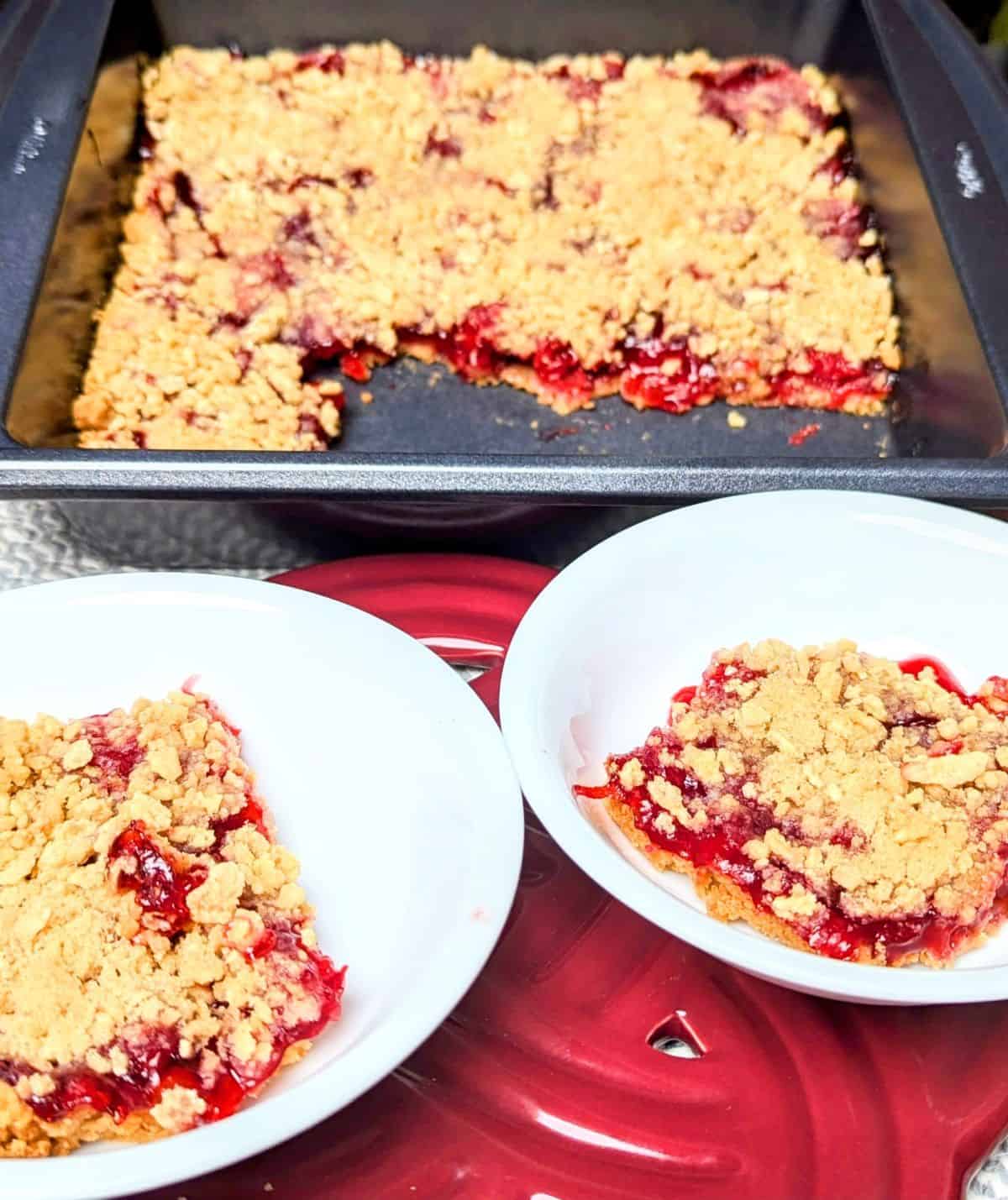 A freshly baked pan of cherry crumble dessert next to two white plates with individual servings resting on a red surface.