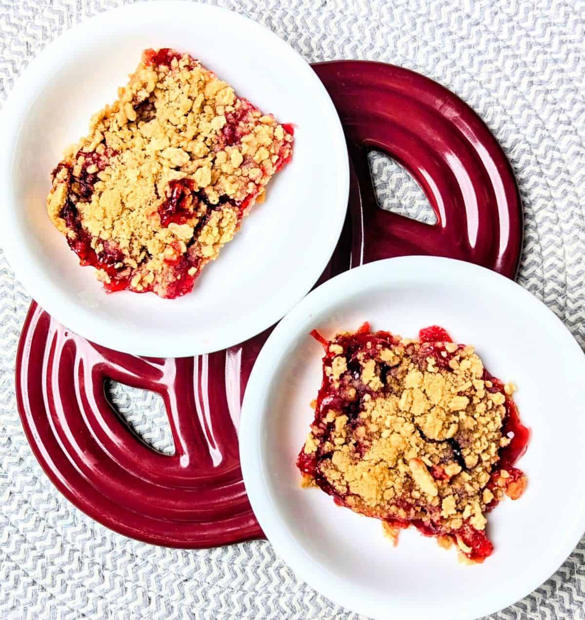 Two square slices of cherry crumble dessert served with vanilla ice cream on white plates over red striped napkins.
