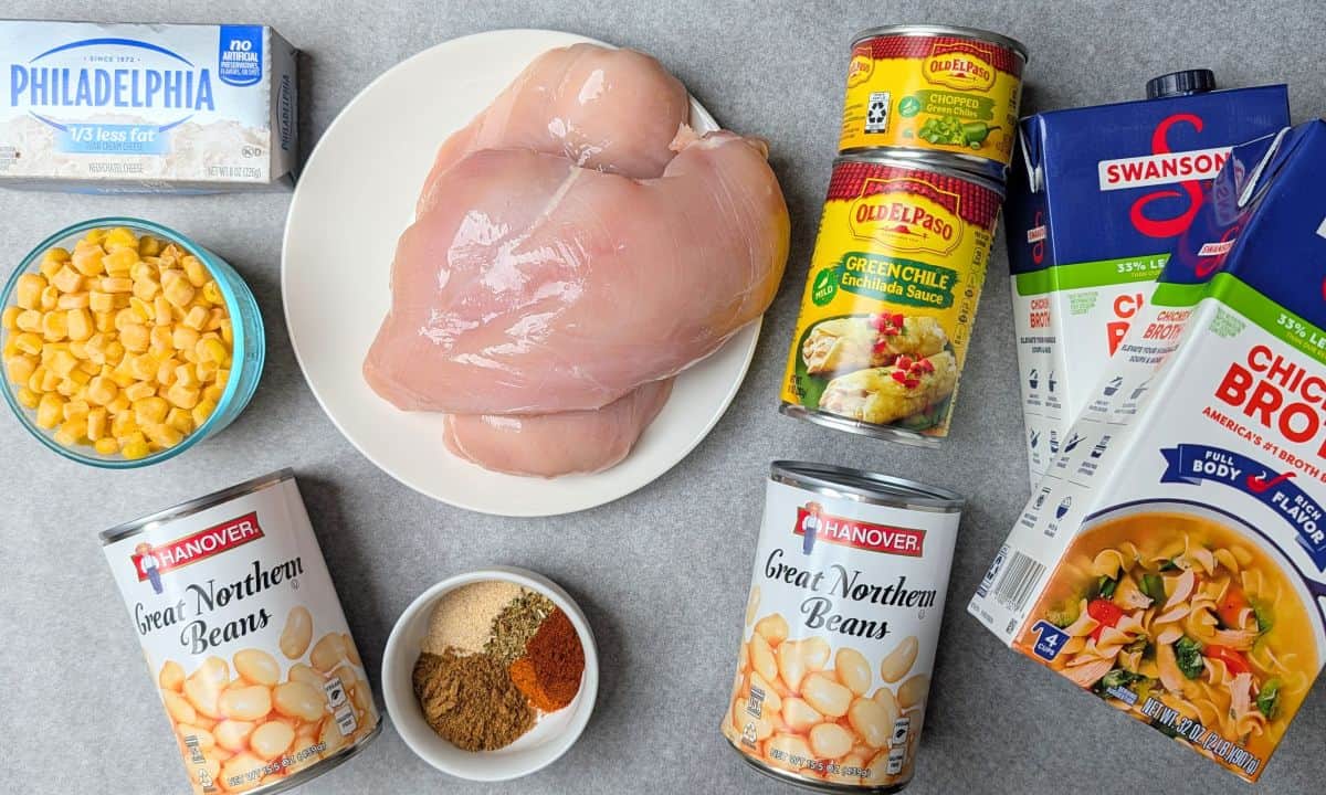 Raw chicken breasts, a block of cream cheese, chicken broth, taco seasoning, and cans of black beans, corn, and diced tomatoes with green chilies arranged on a white surface.