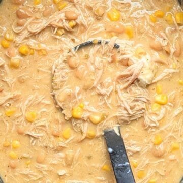 A close-up of a black ladle scooping thick, creamy crockpot cream cheese chicken chili, showing shredded chicken, corn, and beans right out of the slow cooker.