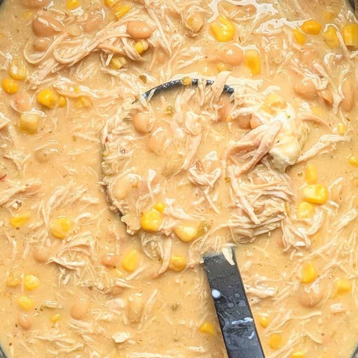 A close-up of a black ladle scooping thick, creamy crockpot cream cheese chicken chili, showing shredded chicken, corn, and beans right out of the slow cooker.