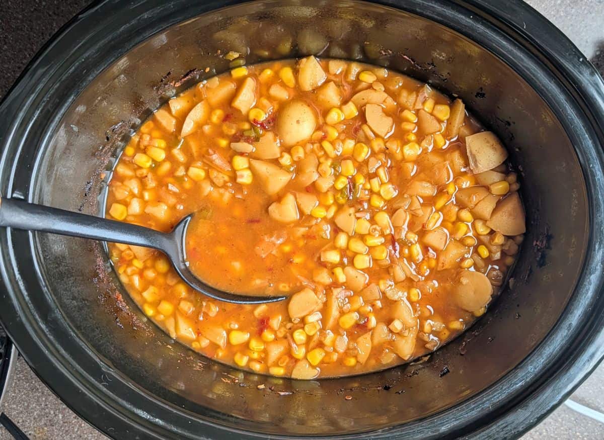 A top-down view of a slow cooker filled with simmered Mexican street corn soup, showing tender corn and potatoes in a rich, warm broth with a black ladle resting inside.