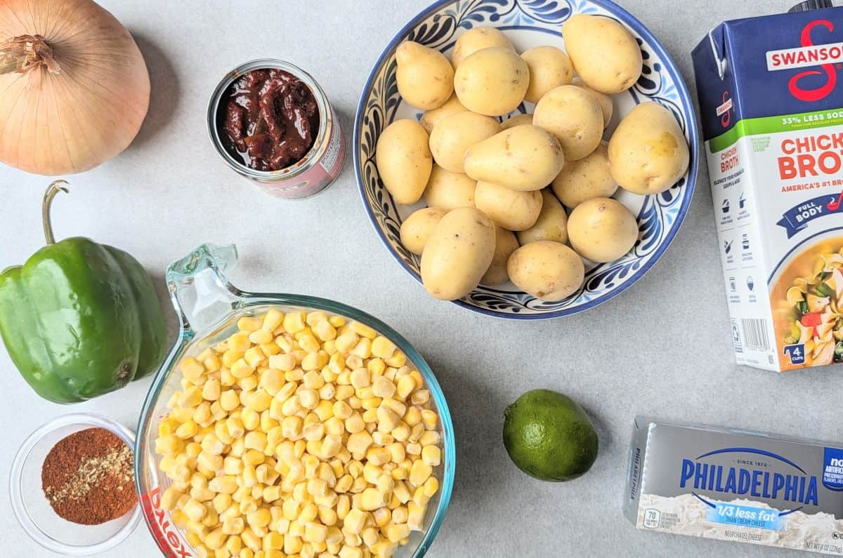 A flat lay of fresh ingredients for creamy Mexican street corn soup, including sweet corn, baby potatoes, a block of cream cheese, an onion, and spices.