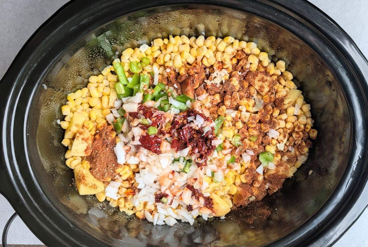 A top-down view of a slow cooker filled with sweet corn kernels, diced onions, and colorful spices, which are the seasonings added to make creamy Mexican street corn soup.