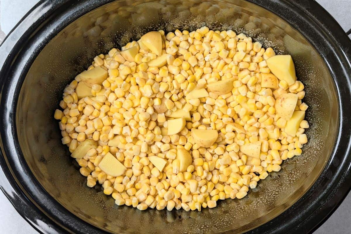 A black slow cooker insert filled with diced potatoes and yellow corn kernels, ready to be cooked for creamy Mexican street corn soup.