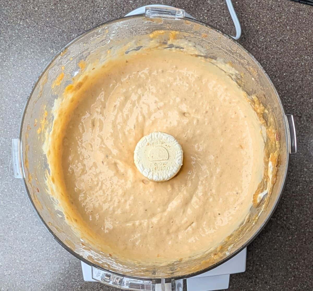 A top-down view of a food processor filled with blended Mexican street corn soup and a block of cream cheese, ready to be mixed into a smooth base.