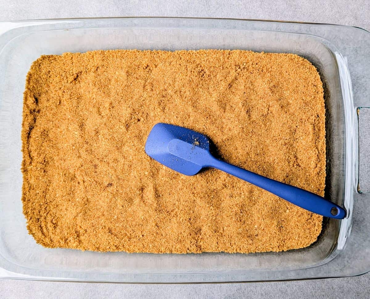 A glass baking dish filled with a prepared graham cracker crust, with a small blue spatula resting inside.