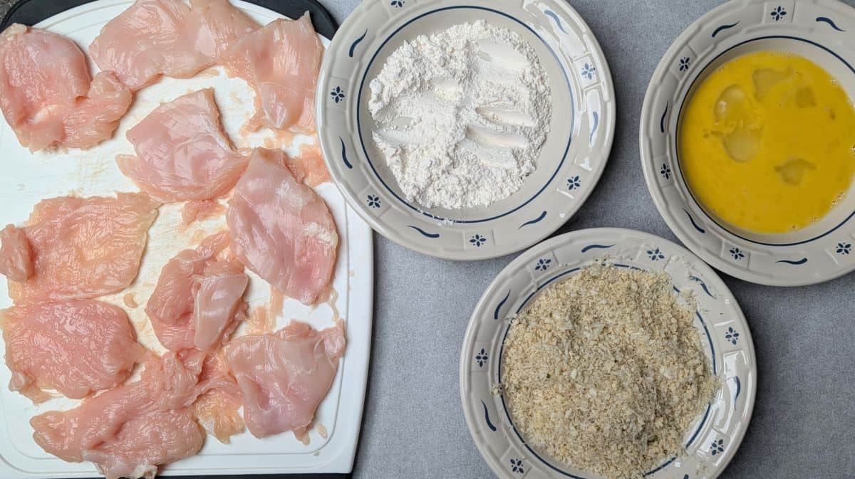 A breading station with bowls of flour, whisked eggs, and panko breadcrumbs next to raw chicken cutlets for healthy chicken parmesan.