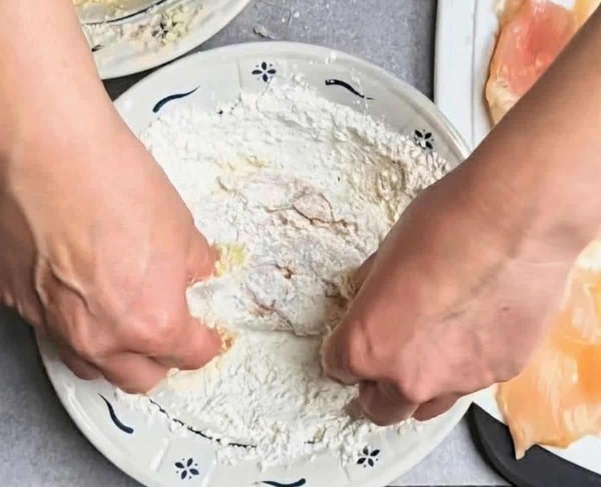 Hands dredging a raw chicken cutlet through a bowl of flour to make healthy chicken parmesan.