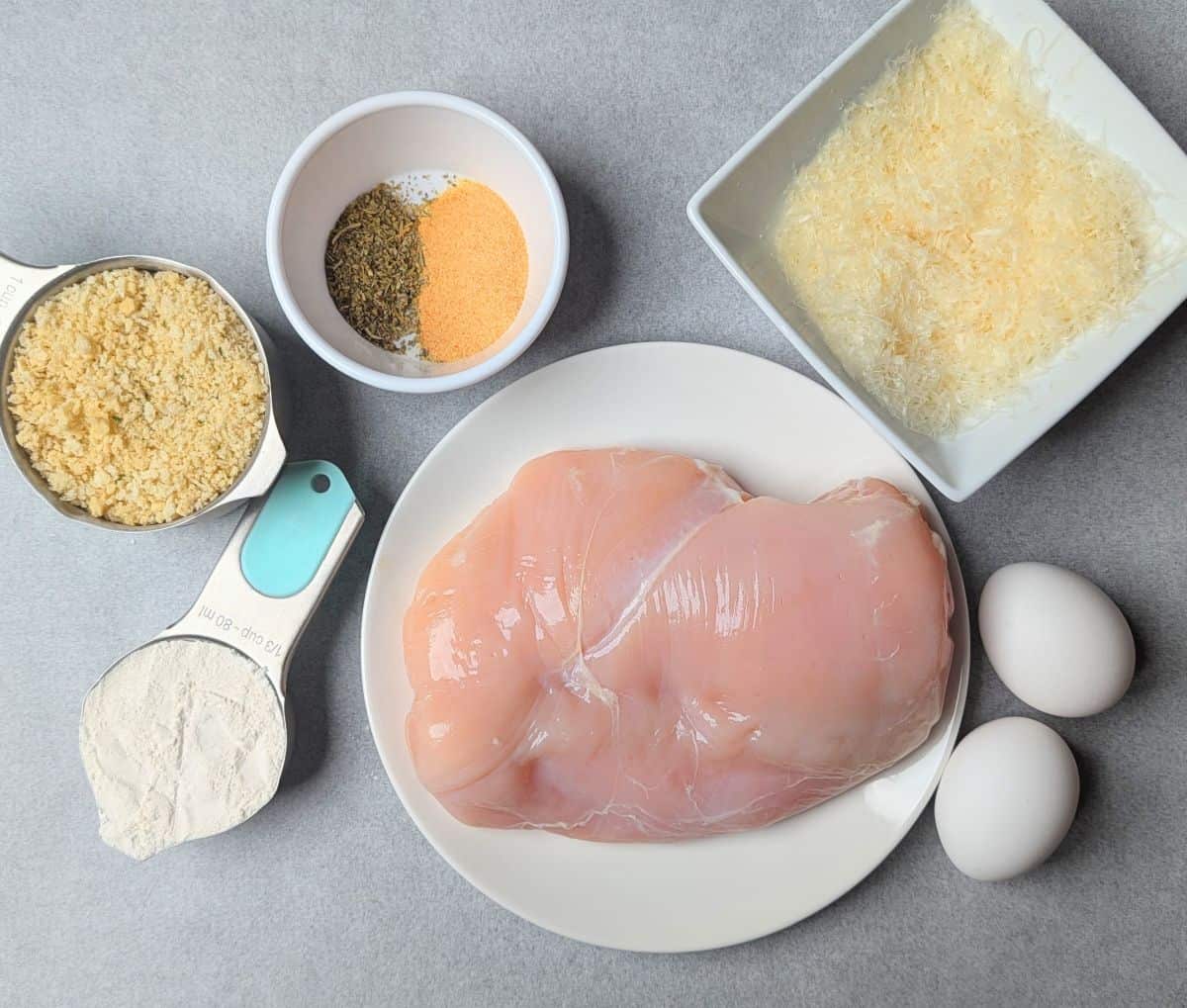 chicken breasts, bowls of breadcrumbs, parmesan cheese, and flour to make healthy chicken parmesan.