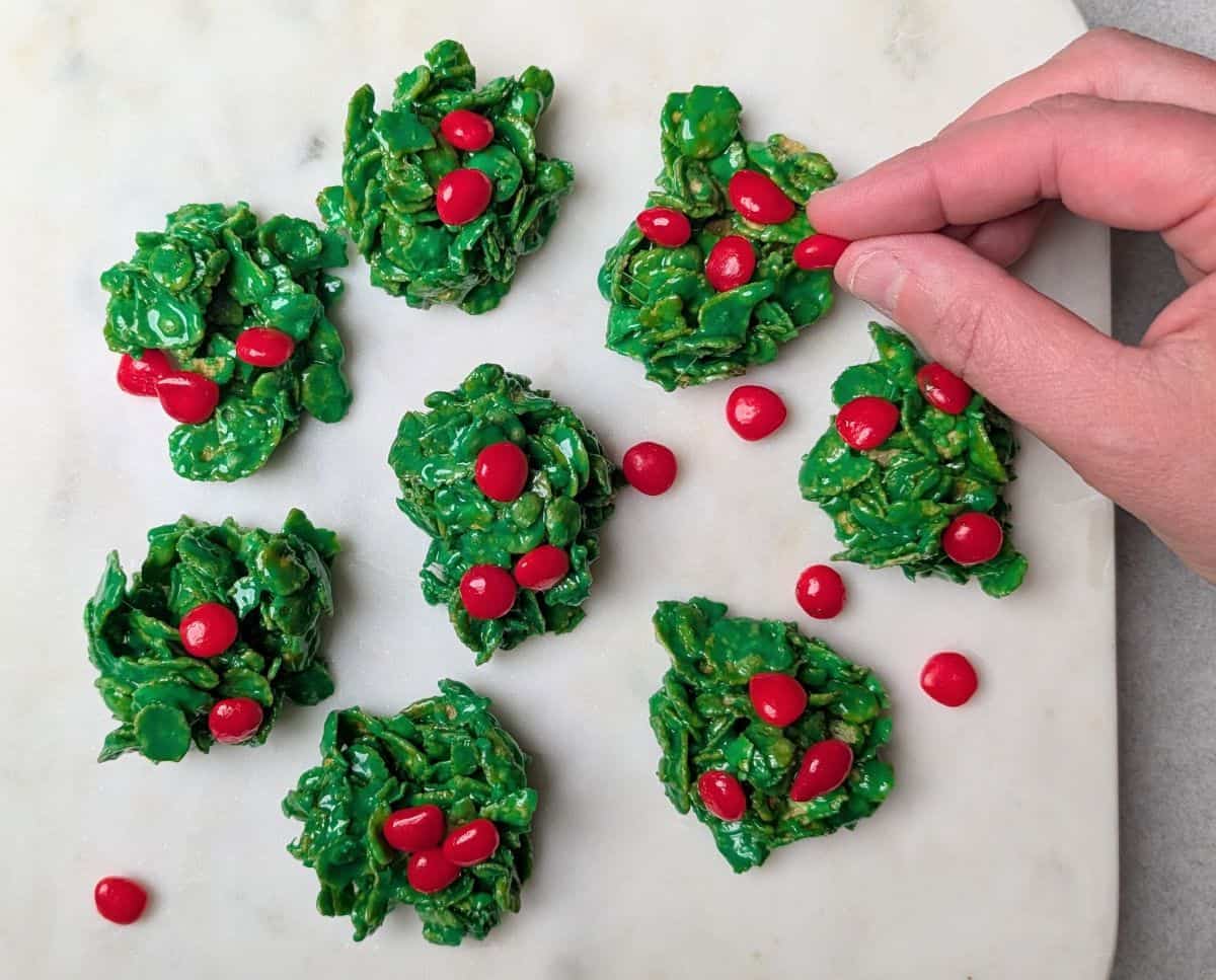 Hands adding small red candy "berries" to the green cookies.