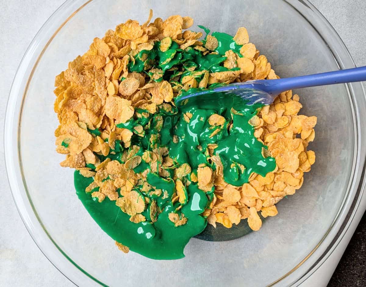 A blue spatula mixing cornflakes into a bright green melted marshmallow mixture.