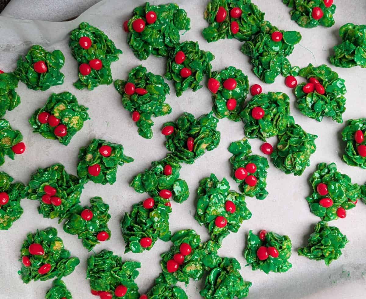 Freshly made no-bake cornflake holly cookies with red cinnamon candies setting on a parchment-lined tray.