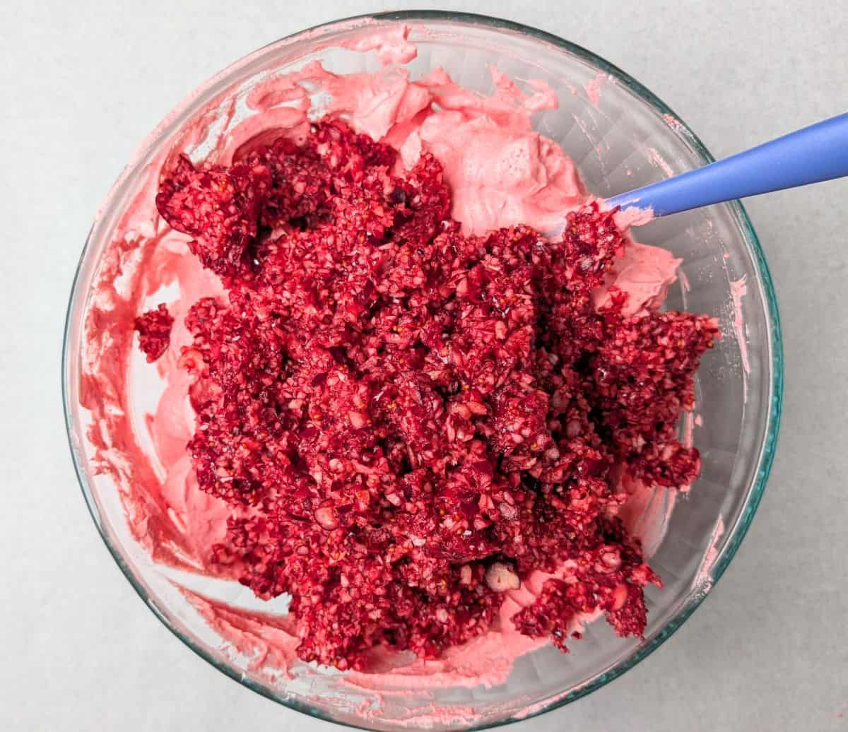 Overhead view of chopped fresh cranberries and lime juice being added to the pink whipped mixture in a clear glass bowl for a cranberry fluff salad recipe.
