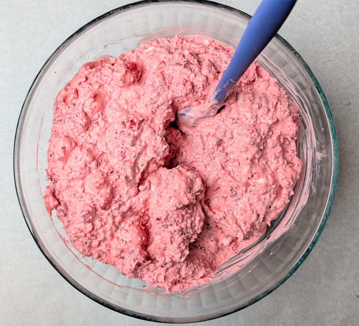 Overhead view of a blue spatula smoothing out the fully combined pink cranberry fluff salad recipe in a clear glass bowl before chilling.