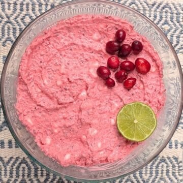 Overhead view of a creamy pink cranberry fluff salad recipe in a clear glass serving bowl, garnished with fresh cranberries and a slice of lime.