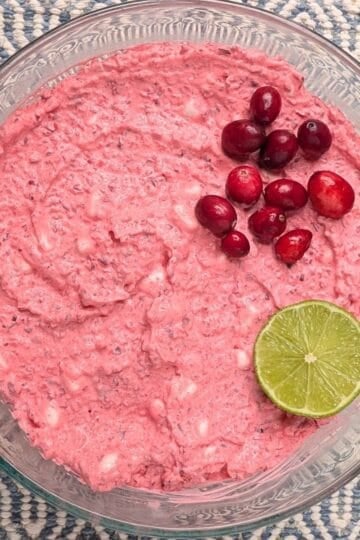Overhead view of a creamy pink cranberry fluff salad recipe in a clear glass serving bowl, garnished with fresh cranberries and a slice of lime.