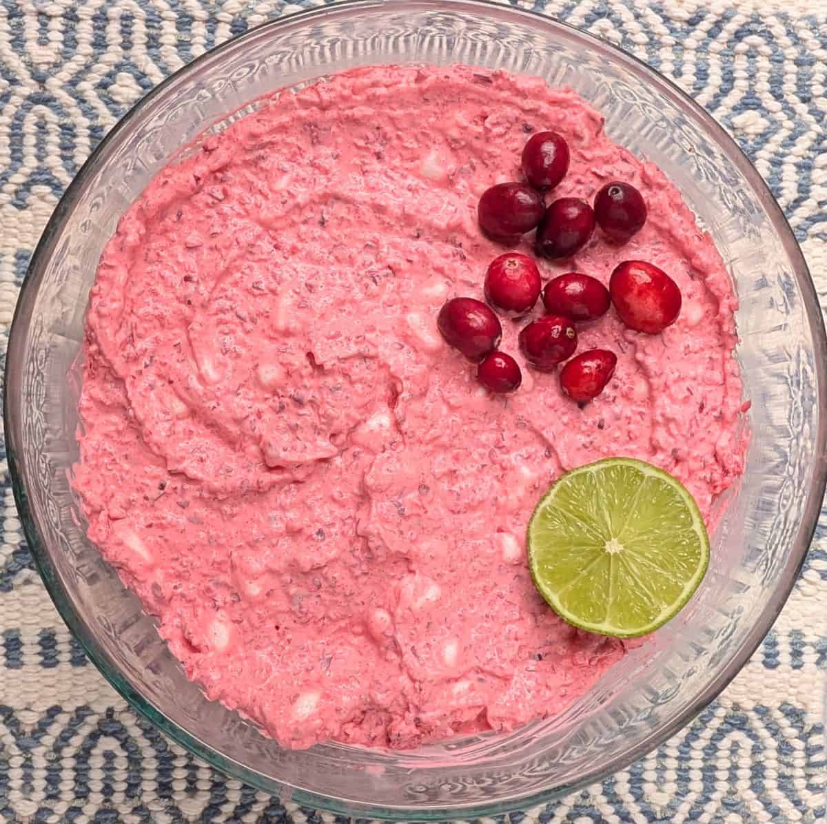 Overhead view of a creamy pink cranberry fluff salad recipe in a clear glass serving bowl, garnished with fresh cranberries and a slice of lime.