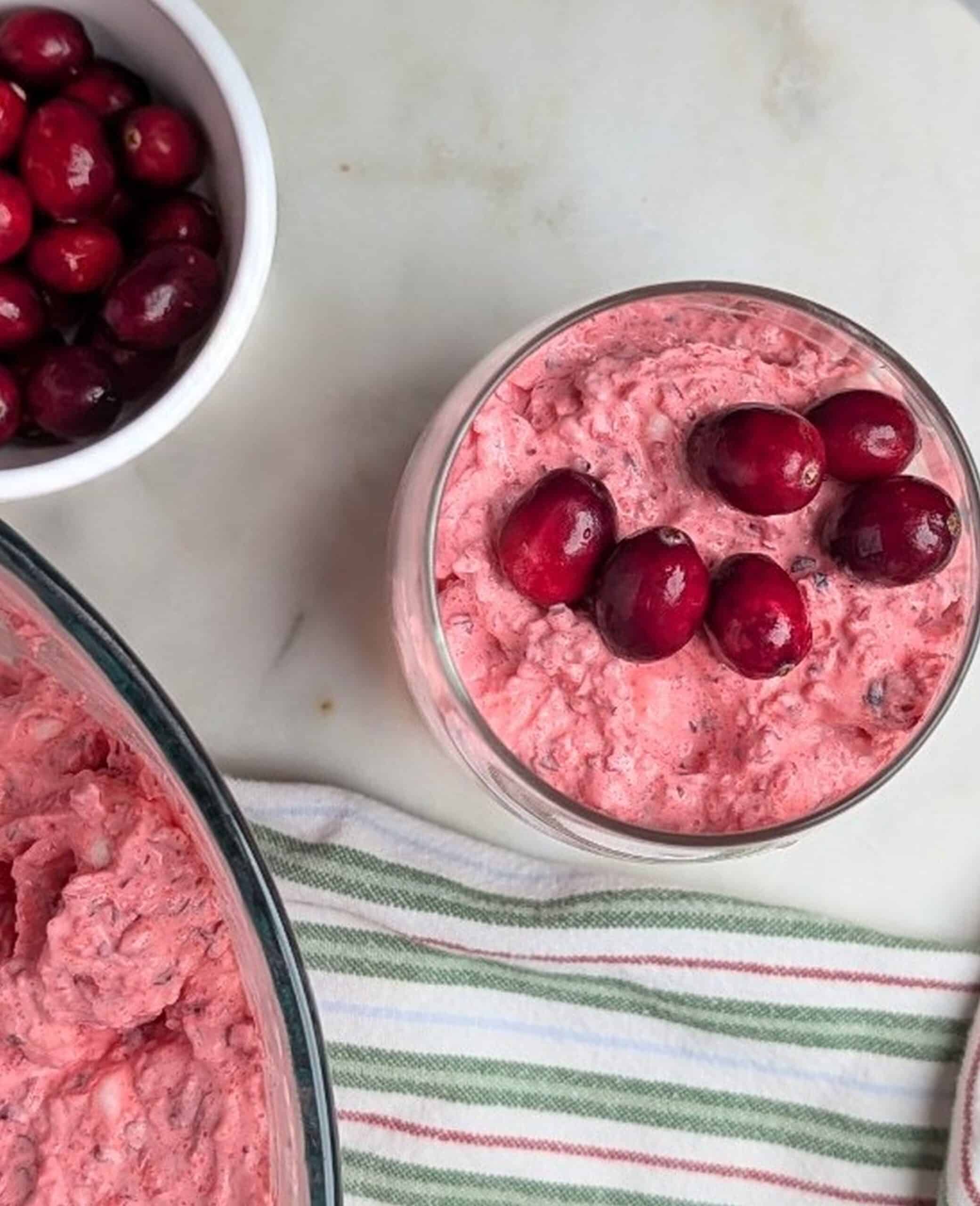 Overhead view of a creamy pink cranberry fluff salad recipe in a clear glass serving bowl, garnished with fresh cranberries on top.