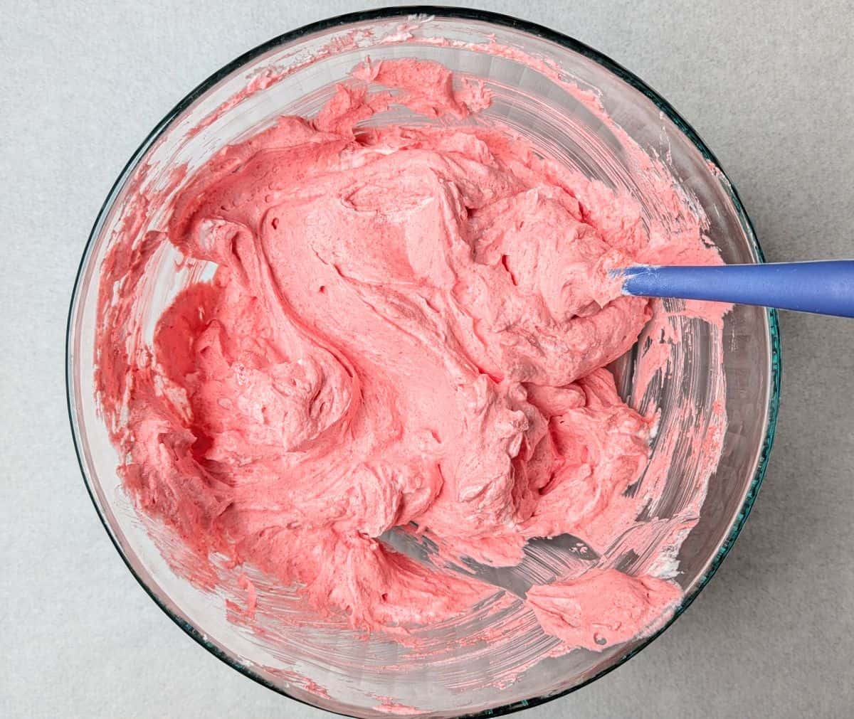 Overhead view of a blue spatula mixing cool whip, Greek yogurt, and raspberry jello together in a clear glass bowl for a cranberry fluff salad recipe.