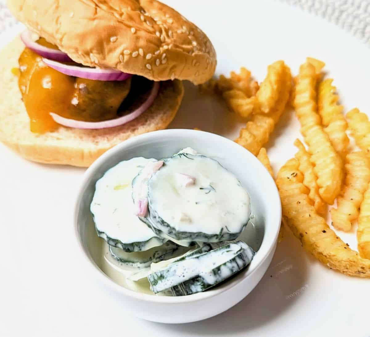 cucumber sour cream salad in serving bowl with hamburger and fries on plate for cookout