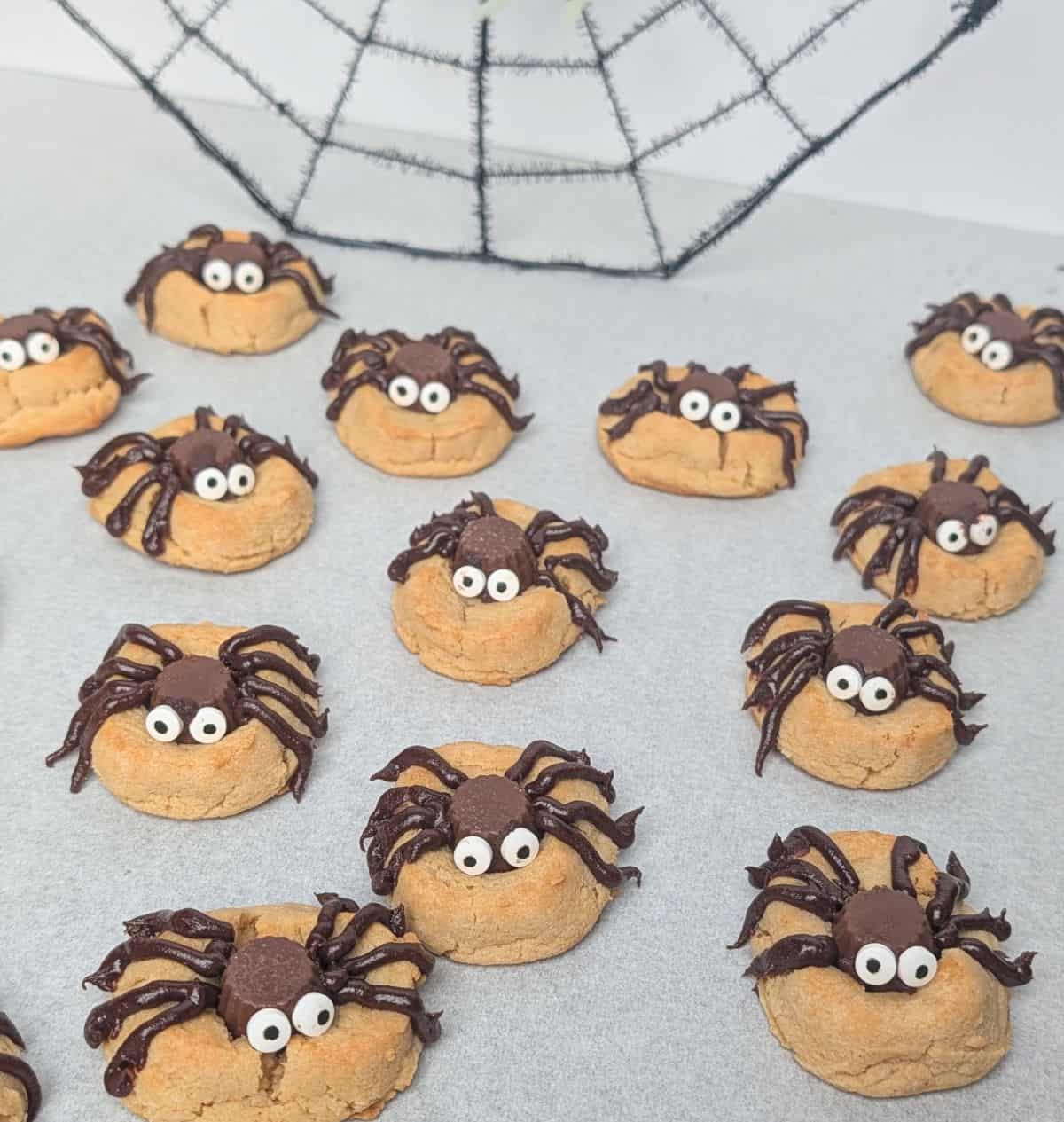 A white serving tray filled with homemade peanut butter Halloween spider cookies decorated with chocolate candy bodies, piped chocolate legs, and candy googly eyes, sitting on a decorative spider web.