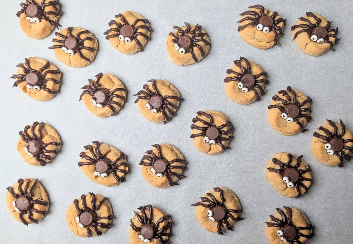 An overhead view of freshly decorated homemade peanut butter Halloween spider cookies with chocolate candy bodies and googly eyes, resting on a parchment-lined baking sheet.