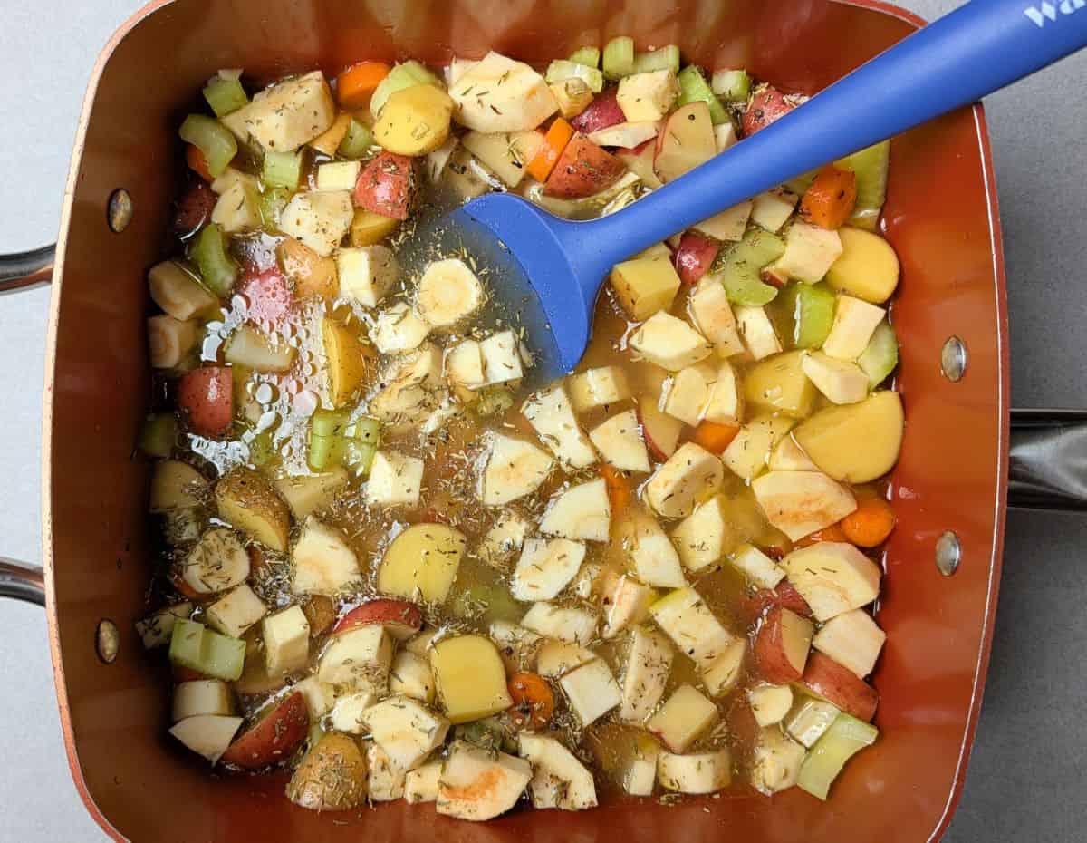 A blue spatula stirring savory broth and seasonings into a mix of chicken, potatoes, and vegetables to create the filling for a healthy chicken pot pie.