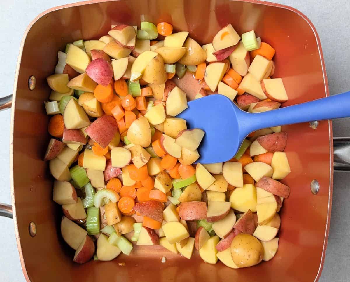 A blue spatula stirring fresh chopped vegetables, including carrots, potatoes, and onions, in a pot to make the filling for a healthy chicken pot pie.
