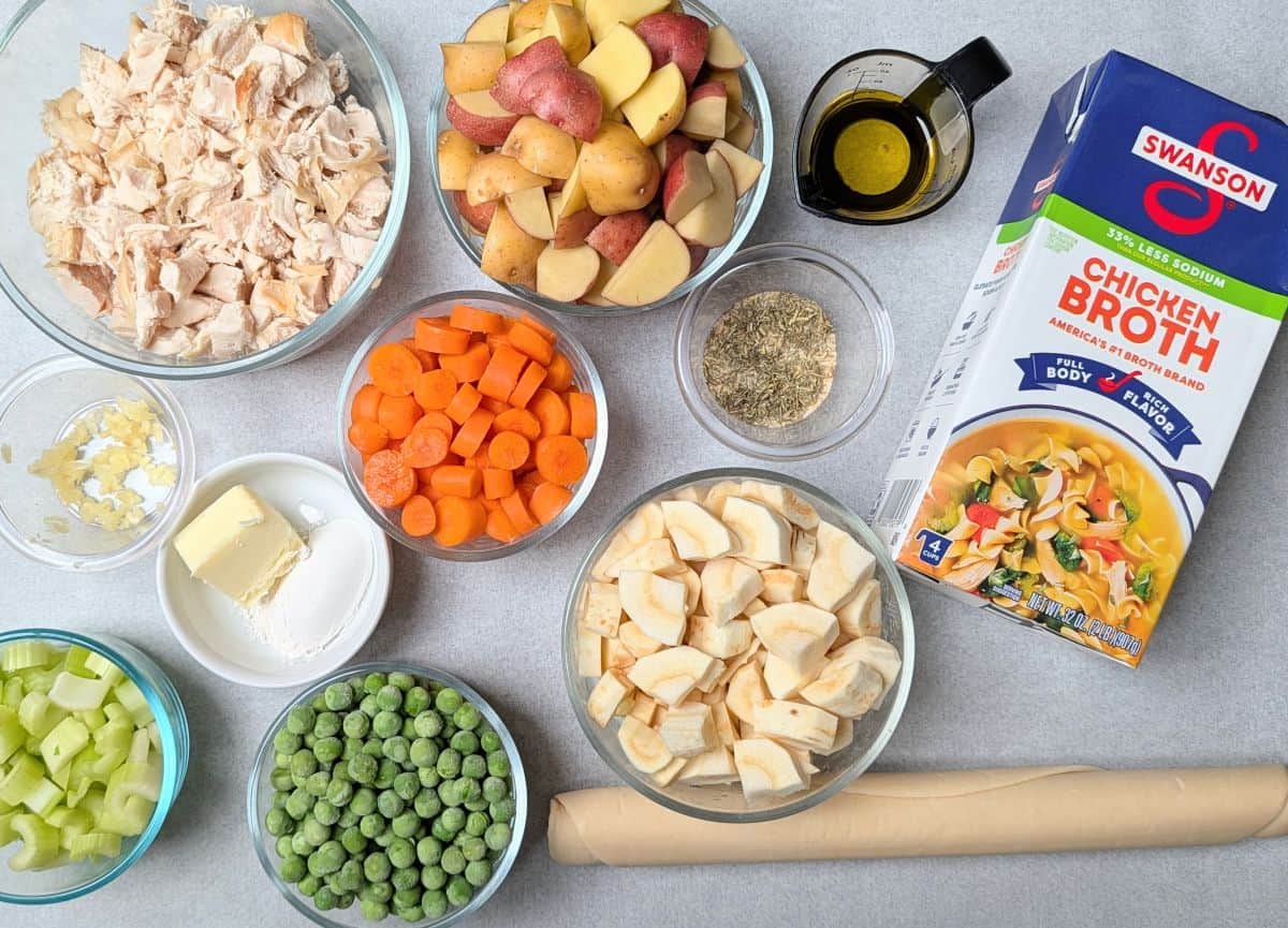 Prepped ingredients for healthy chicken pot pie, including small bowls of diced chicken, carrots, peas, potatoes, and a carton of broth.