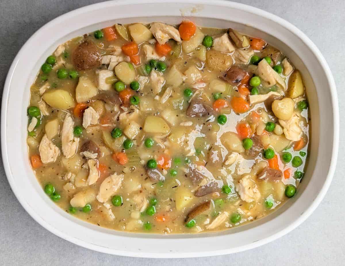 A white casserole dish filled with healthy chicken pot pie filling, showing a savory mix of diced chicken, peas, carrots, and potatoes ready for the crust.