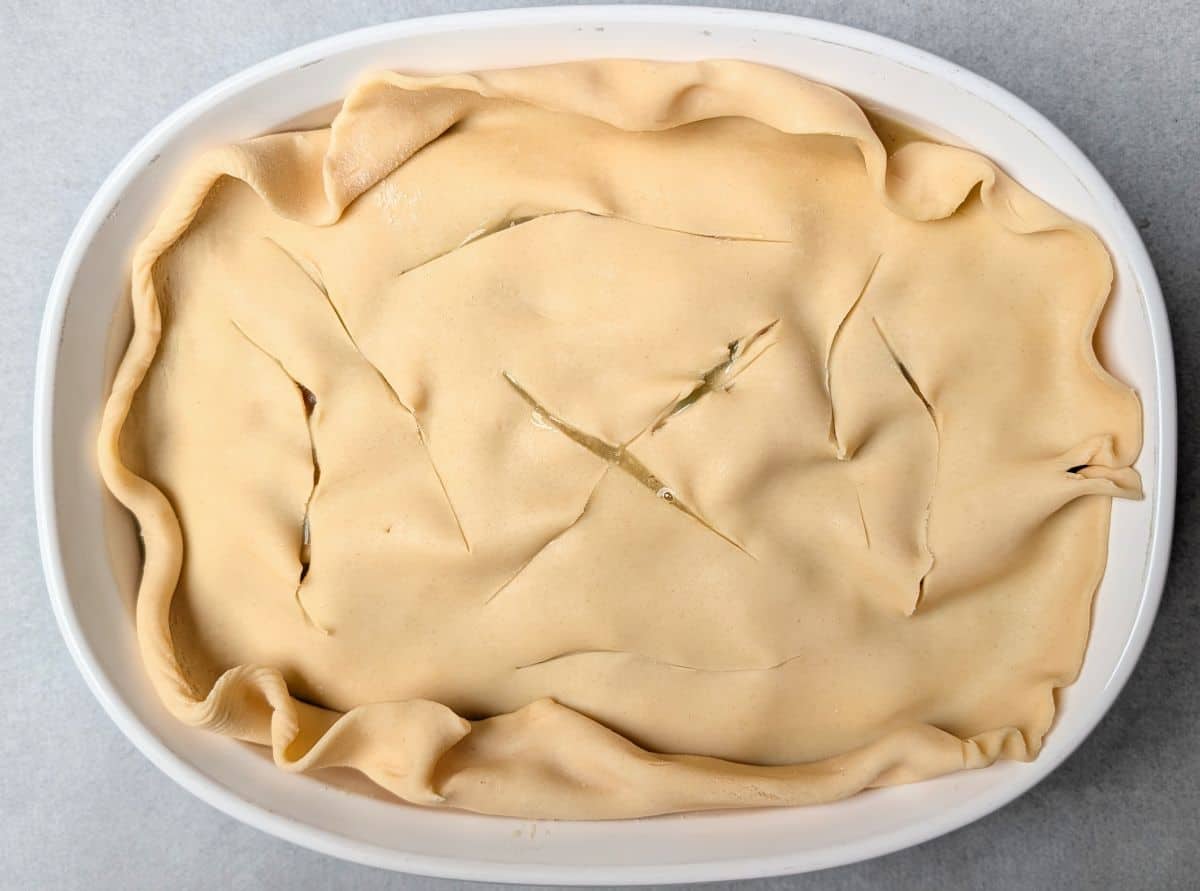A raw pie crust placed over a casserole dish filled with healthy chicken pot pie, showing several small slits cut into the dough to vent steam before baking.