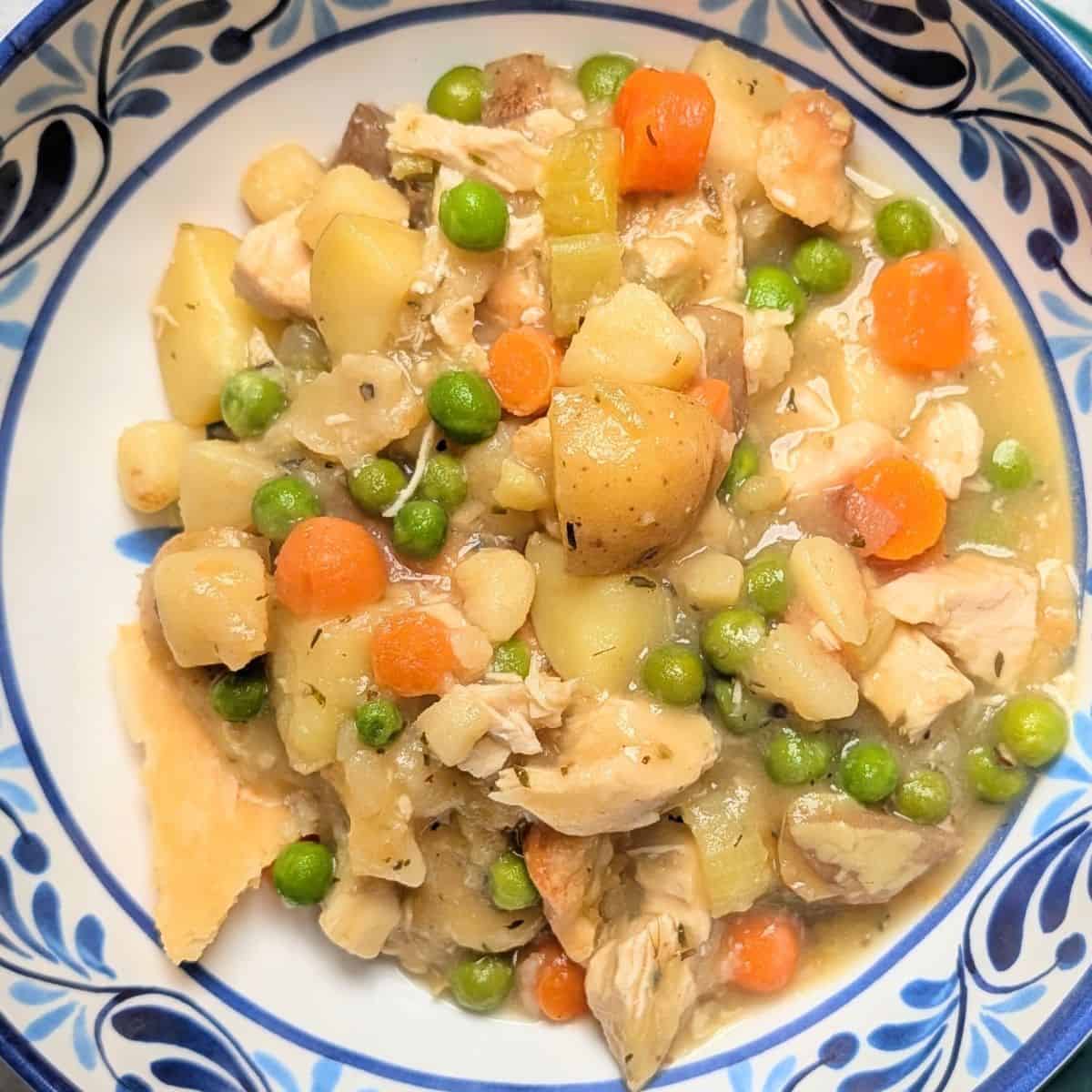 A serving of healthy chicken pot pie in a blue patterned bowl, showing tender chunks of chicken, peas, carrots, and potatoes, topped with pieces of golden baked pie crust.
