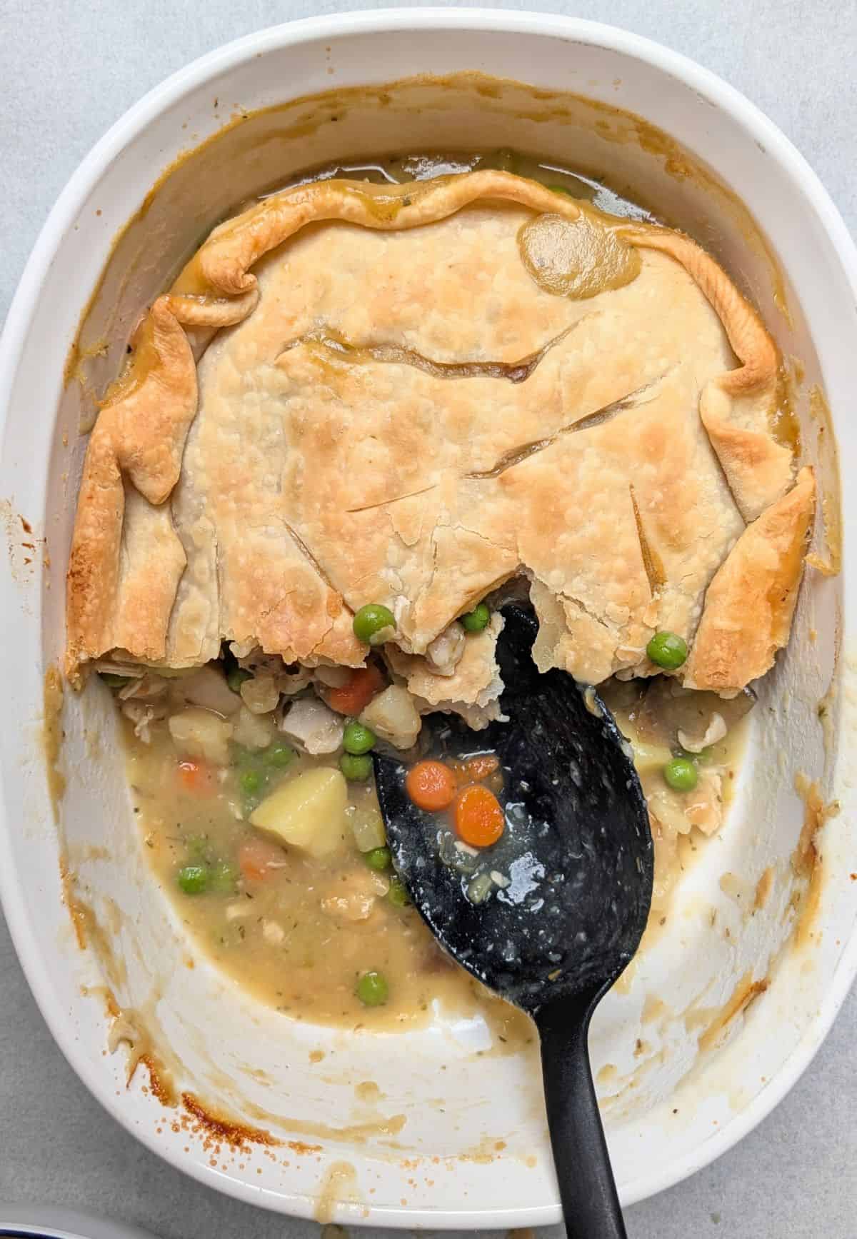 A black serving spoon scooping up the filling of a healthy chicken pot pie from a casserole dish, showing tender pieces of chicken, peas, carrots, and savory broth