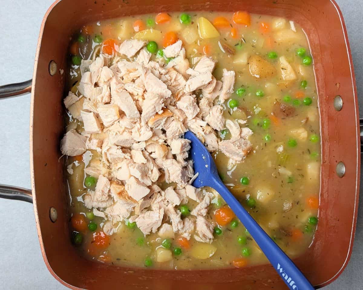 A blue spatula stirring chunks of leftover skinless chicken breast into a mix of vegetables and broth to make the filling for a healthy chicken pot pie.